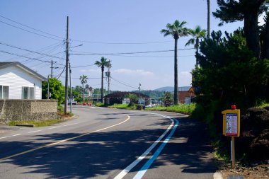Mavi bisiklet şeridine sahip bir köy yolu Taeheung yakınlarındaki evler, palmiye ağaçları ve elektrik hatlarının yanından geçiyor. Tepeler ve inşaat varilleri uzaktan görülebiliyor..