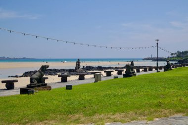 Bir sıra taş tabure ve sahil hayvan heykelleri Pyoseon Sahili 'nde sığ göle ve açık denize bakan çimenli gezinti güvertesinde sıralanır. Ziyaretçiler uzak kumsalda duruyor.