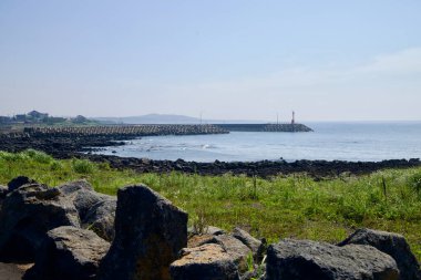 Beton zırh bloklarıyla kaplı uzun bir dalgakıran, Taeheung limanını korur, kırmızı bir fenerde, siyah lav kayası ve ön planda durgun su ile sona erer..