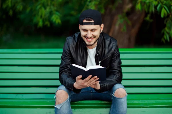 Jovem bonito lendo um livro e sorrindo no parque . — Fotografia de Stock