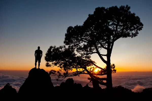 Man silhouette with hands in pockets stand on the rock peak and watching the sunset with beautiful clouds.