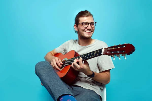 Młody przystojny z okularów grając na gitarze siedząc na białym tle niebieski. — Zdjęcie stockowe