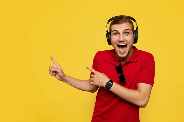 Joven barbudo con polo rojo y auriculares apuntando hacia afuera y sonriendo sobre fondo amarillo . — Foto de Stock