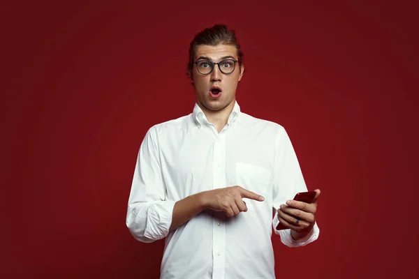 Retrato de un joven sorprendido con camisa blanca y gafas apuntando con el dedo al teléfono móvil aislado sobre fondo rojo — Foto de Stock