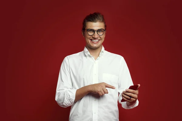 Χαρούμενος νεαρός άνδρας στο λευκό πουκάμισο και τα γυαλιά, χαμογελώντας και δείχνοντας το δάχτυλό κινητό τηλέφωνο που απομονώνονται σε κόκκινο φόντο — Φωτογραφία Αρχείου