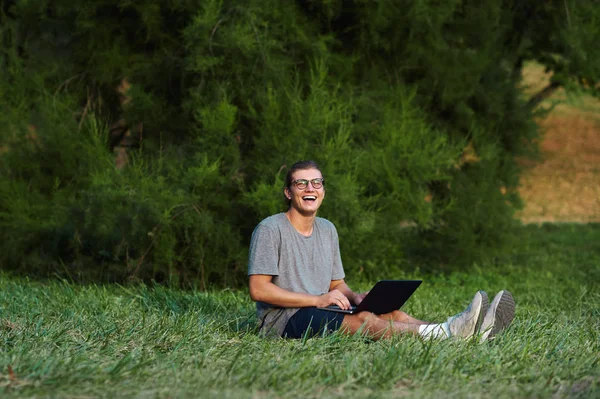 Dizüstü holding ve genel Park çimenlerin üzerinde otururken kameraya gülme neşeli delikanlı. Açık eğleniyor öğrenci.