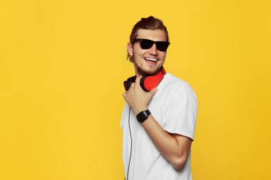 Neşeli hipster adam gülümsüyor ne mutlu ki, Beyaz tişörtlü, güneş gözlüğü ve sarı üzerinde izole kulaklık, ifade heyecan