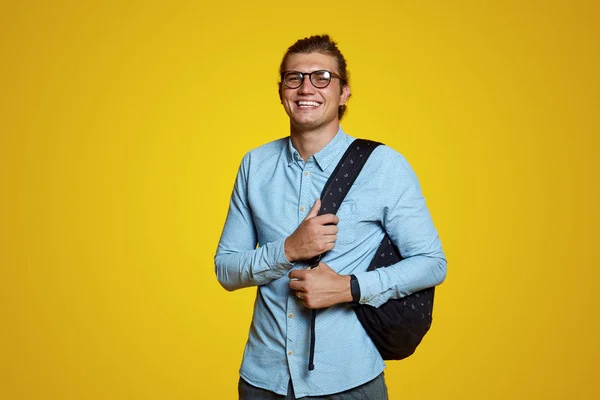 Atractivo chico joven en camisa azul y gafas con mochila y sonriendo a la cámara en el fondo amarillo — Foto de Stock