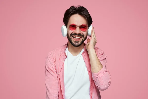 Attractive bearded male listens favourite song, likes beats in headphones, connected to modern cellular, isolated on pink backdrop — Stock Photo, Image