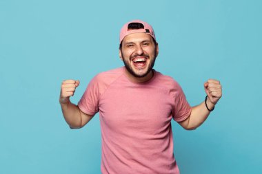 Neşeli genç çocuk yumruklarını clenches, mutluluktan bağırıyor, zafer kutluyor, kap ve üzerinde mavi duvar izole pembe tişörtü giyer