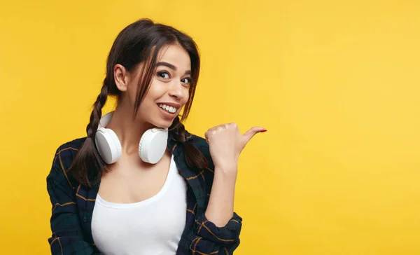 Sağ tarafta, çekici genç bayan Puan rahat gömlek ve beyaz kulaklık giyen, sarı arka plana karşı boş alan gösterir