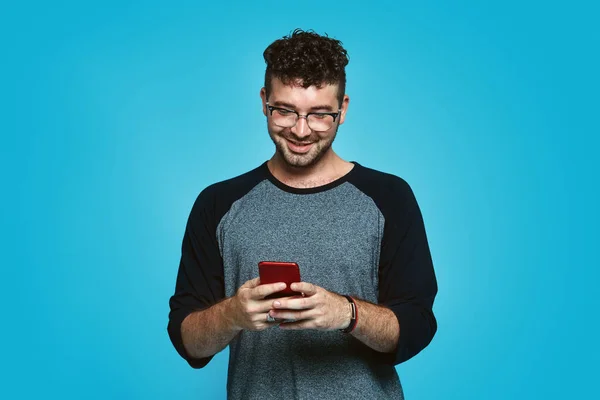 Imagen del hombre barbudo sonriente con anteojos escribiendo mensaje en el teléfono inteligente sobre fondo azul — Foto de Stock