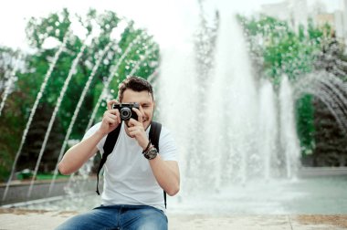 Yakışıklı genç adam parkta fotoğraf çekiyor. Beyaz bir t-shirt şık çocuk, çanta ile inanılmaz çeşme önünde fotoğraf çekmek