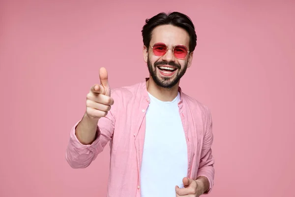 Çekici adam, pembe gömlek ve güneş gözlüğü giyiyor, gülümseyen ve kamera işaret, pembe duvara karşı poz.