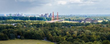 Duisburg panorama bridges Rhine River industrial skyline. Scenic wide-angle view over green forest landscape with two bridges crossing the Rhine and factories in the background, showing contrast between nature and urban infrastructure