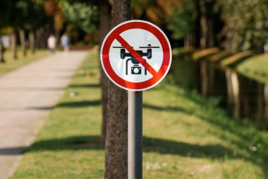 No Drones Sign Park Pathway Trees Summer. Close-up of a red prohibition sign forbidding drone use in a public park area with a walking path, green grass, and trees. Emphasizes safety, privacy, and regulation in recreational spaces
