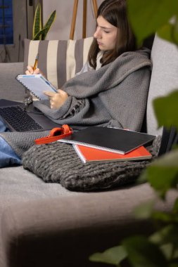 Blonde adolescent using laptop in living room, remote work lifestyle, young freelancer or student working from home. Modern cozy interior. Digital education lifestyle concept