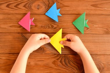 Origami renkli balıklar. Çocuk elinde sarı bir origami balığı tutuyor. Kahverengi ahşap arka plan. Origami kağıt işi fikri. Kolay kağıt işçiliği. DIY origami balığı nasıl yapılır. Çocuklar için balıkçı tekneleri. Çocuklar için kağıt balık origamisi