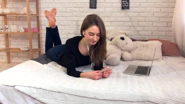 漂亮的女人听音乐智能手机与耳机在她的卧室 女孩使用手机和放松床 — 图库视频影像