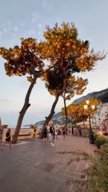 Yerel halk ve ziyaretçiler Amalfi Sahili boyunca sakin bir yürüyüşün tadını çıkarırken, sıcak akşam ışığı süslenmiş ağaçlara parlıyor. Manzaralı görüntüler şenlikli atmosferi artırır.