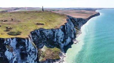 Experience the stunning aerial perspective of Cap Blanc Nez in France. This location features dramatic white cliffs meeting the sparkling sea, surrounded by lush green fields under a clear sky.