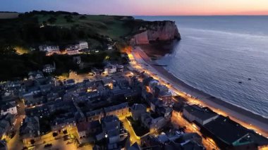 Experience the tranquil beauty of Etretat as the evening unfolds. The coastal town glows with soft lights while the cliffs and ocean reflect the setting sun.