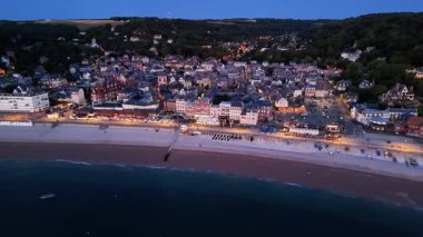 Explore the evening charm of Etretat with a stunning aerial perspective over the coastline and town. Observe the vibrant lights that illuminate the streets as night falls.