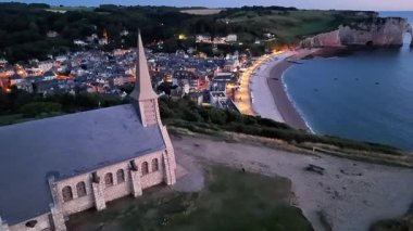 Experience Etretat in the evening as aerial views reveal stunning white cliffs, twinkling village lights, and a peaceful coastal setting by the sea. The charming landscape enchants visitors.
