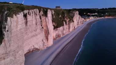 Experience the breathtaking cliffs of Etretat as the sun sets, casting a warm glow over the beach and illuminating the charming village along the shoreline.