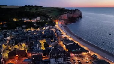 Experience a breathtaking evening aerial view of Etretat, France, where the charming coastal town and iconic cliffs are beautifully lit against the twilight sky.