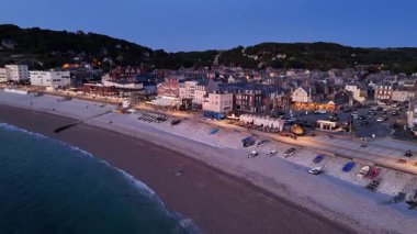 Experience an evening over Etretat beach in France, where the calm waves meet the soft glow of lights along the shore. The charming seaside town comes alive at dusk, creating a tranquil atmosphere.