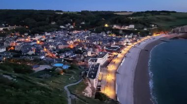 Experience the tranquil beauty of Etretat as the evening sun sets, casting a warm glow over the seaside town. Streets and beachfront come alive with lights, creating a picturesque view.