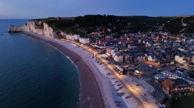 Majestic cliffs of Etretat rise above the serene beach as evening settles in. Charming coastal town lights up, offering a warm welcome to visitors. Discover the beauty of this enchanting landscape.