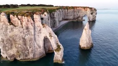 Experience the breathtaking aerial view of the Etretat cliffs in France. Marvel at the iconic rock formations, lush greenery, and serene waters of the coastline during golden hour.