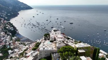 Experience a stunning aerial view of Positano, Italy. The vibrant buildings cascade down the cliffs, overlooking the beautiful blue waters and boats dotting the coastline.