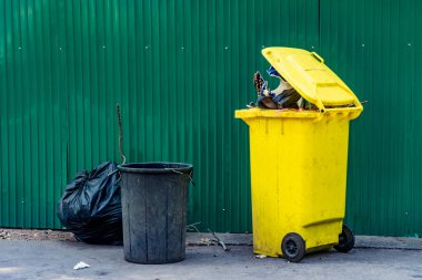 Sarı ve siyah plastik çöp tenekesi. Beton yolda yeşil metal çitli.