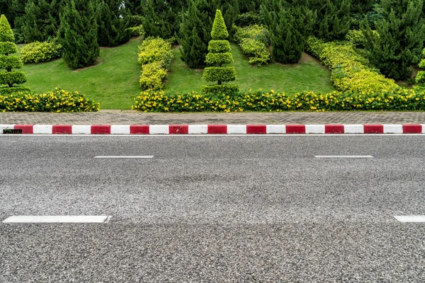 Asfalt yolu, kırmızı ve beyaz beton kaldırım ve küçük yeşil çimen tepesinde çam ağaçları.