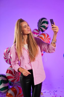 Kulaklıklı, akıllı telefonlu bir kız, bir şarkı söylüyor, mor renkli balonlarla, şekerlerin arka planında