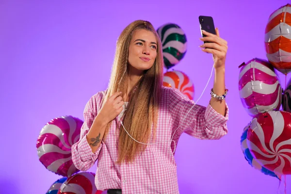 Girl in headphones with a smartphone, sings a song, on a purple background of candy, colored balloons