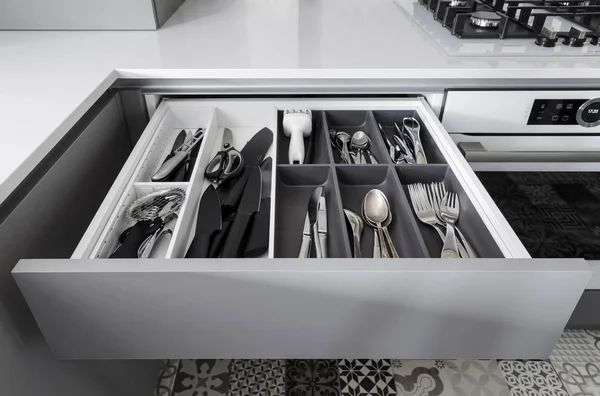 Opened kitchen drawer with a tray and cutlery inside. Grey front of the drawer, white inside, handless design.