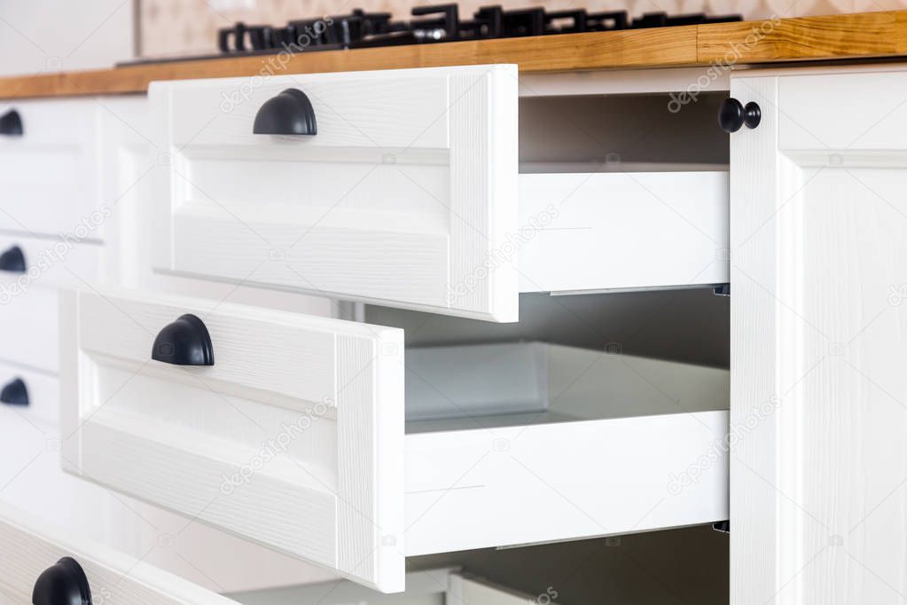 Opened kitchen drawers, kitchen in a traditional style with wooden white facade, black handles and wooden countertop 