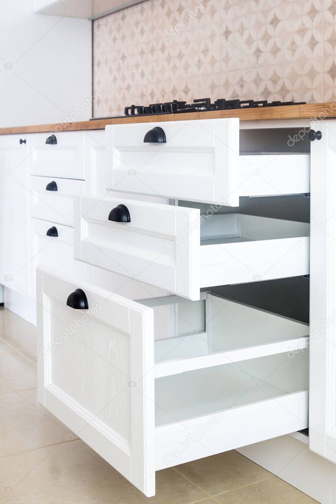 Opened kitchen drawers, kitchen in a traditional style with wooden white facade, black handles and wooden countertop 