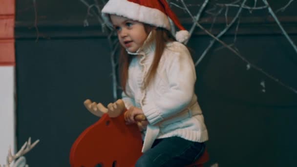 スローモーション小さな女の子は装飾されたクリスマスツリーの近くにロッキング馬の上に座ります — ストック動画