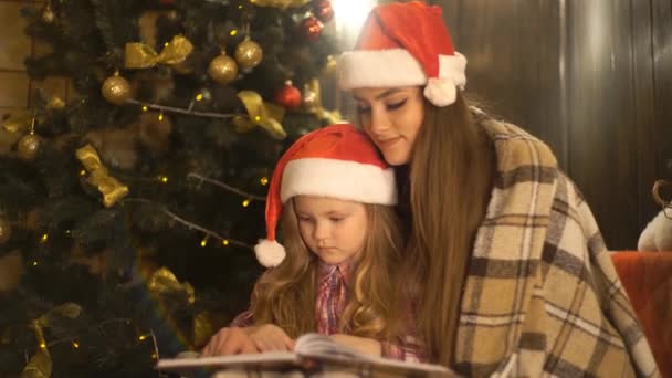 戴着圣诞老人帽子的女孩在圣诞树前看书 — 图库视频影像