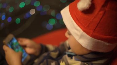 Noel Baba Şapkası Giyen Genç Çocuk Noel ağacının yanında cep telefonuyla oyun uygulaması oynuyor..