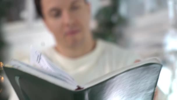 Νεαρός Άνδρας Διαβάζει Δυνατά Από Ένα Βιβλίο Δίπλα Στο Χριστουγεννιάτικο — Αρχείο Βίντεο