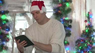 Bir adam Noel ağacının yanındaki sandalyede oturur ve kitap okur.