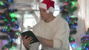 Bir adam Noel ağacının yanındaki sandalyede oturur ve kitap okur.