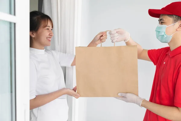 Asian delivery man holding shopping bag in medical rubber gloves and mask give to young woman in front of the house,