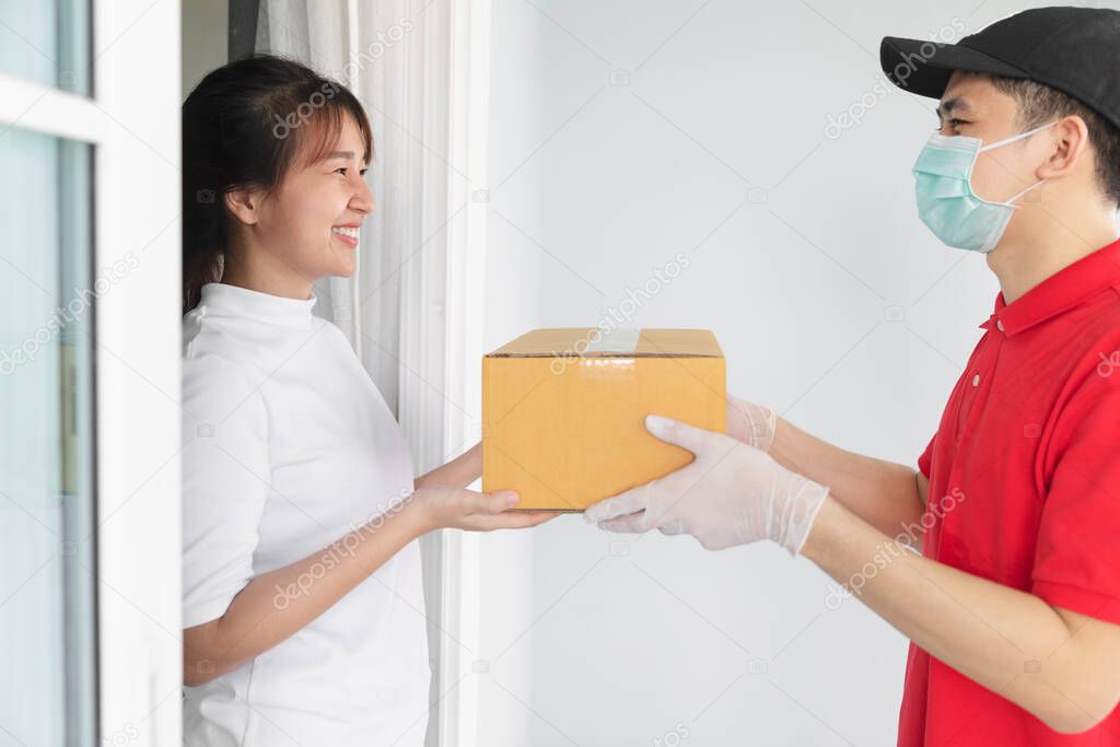 Asian delivery man holding cardboard boxes in medical rubber gloves and mask give to young woman in front of the house,